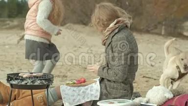 母亲带着女儿，一个穿着外套的年轻女人坐在河边的海滩上，大海，野餐，<strong>做饭</strong>，<strong>做饭</strong>。